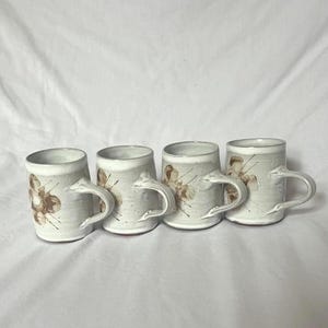 May include: Four handmade ceramic mugs. Each mug is off-white with a brown floral design and a curved handle. The mugs are cylindrical and appear to be of similar size and shape, perfect for coffee or tea.