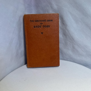 Könnte beinhalten: Ein kleines, rechteckiges Buch mit braunem Einband. Der Titel "THE OBSERVER'S BOOK OF BIRDS' EGGS" ist in Schwarz auf der Vorderseite aufgedruckt. Das Buch steht auf einer weißen Oberfläche, mit einem hellblauen Hintergrund.