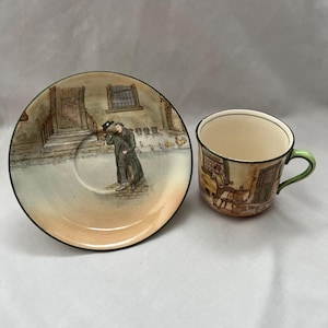 May include: A vintage teacup and saucer set. The saucer depicts a painted scene of a man in a long coat on a street. The teacup has a green handle and a matching scene. The set has a cream interior and a black trim.