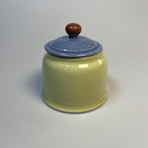 May include: A ceramic jar with a lid. The jar is a light yellow color with a speckled texture. The lid is blue and has a brown knob on top. The jar is round and has a simple, classic design.