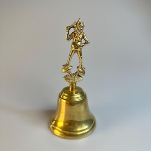 May include: A vintage brass bell with a detailed figurine of a mischievous elf atop. The bell has a classic shape with a flared base and a small clapper inside. The elf is posed with one hand on its hip and the other holding a small object.