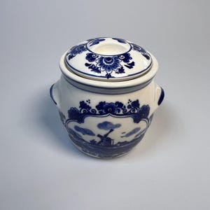 May include: A white ceramic jar with a lid, adorned with blue floral and landscape patterns. The jar has two small handles and a matching lid with a central floral design. The landscape depicts a windmill and clouds.