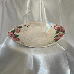 May include: Oval-shaped, off-white ceramic serving dish with a floral design. The dish features a spiral pattern in the centre and is decorated with pink flowers and green leaves on each end. The dish has a pearlescent sheen.