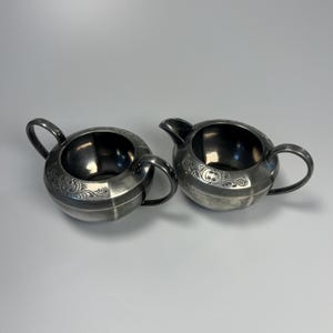 May include: A silver-colored creamer and sugar bowl set. The creamer has a spout and a handle, while the sugar bowl has two handles. Both pieces have a round shape and decorative floral patterns. The set is made of metal.