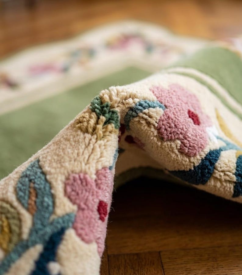 May include: A tufted rug featuring a solid sage green centre panel bordered by a cream-coloured edge. The border is decorated with a repeating floral pattern in shades of dusty pink, dark blue, and olive green, showcasing a thick, textured pile.