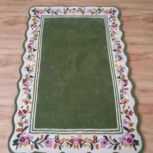 May include: A rectangular rug with a solid olive green centre and a cream-coloured border featuring a scalloped edge. The border is decorated with a repeating floral pattern in shades of pink, yellow, and dark green, framing the central area.
