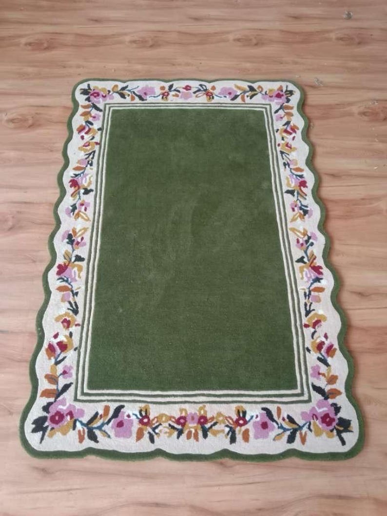 May include: A rectangular rug with a solid olive green centre and a cream-coloured border featuring a scalloped edge. The border is decorated with a repeating floral pattern in shades of pink, yellow, and dark green, framing the central area.