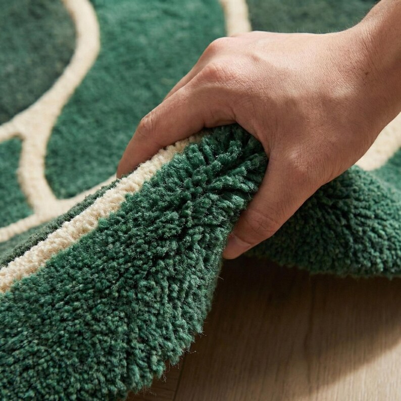 May include: A green and cream-colored rug with a textured, plush surface. The rug features a pattern of interconnected cream-colored shapes against a green background. The rug is being held up, revealing its thickness and construction.