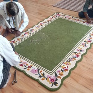 May include: A rectangular green rug with a scalloped edge and a cream-coloured border featuring a colourful floral pattern of pink, orange, and blue flowers with green leaves. The rug has a solid green centre and is being trimmed with scissors.