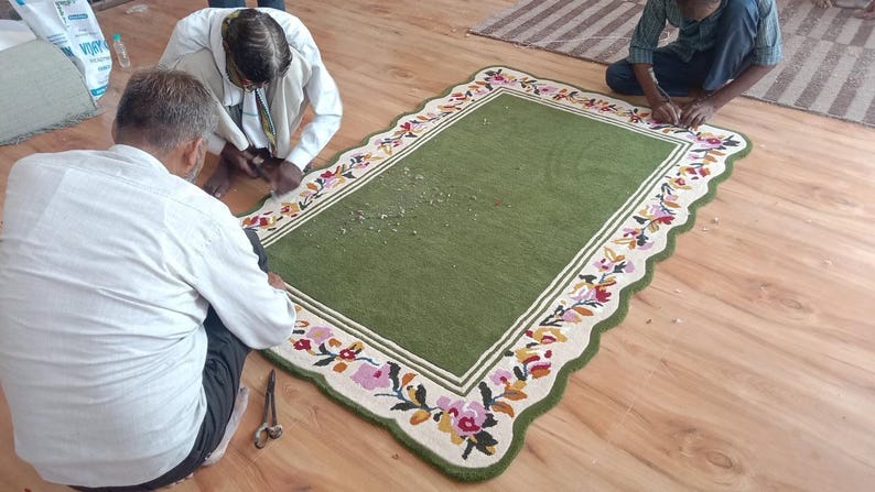 May include: A rectangular green rug with a scalloped edge and a cream-coloured border featuring a colourful floral pattern of pink, orange, and blue flowers with green leaves. The rug has a solid green centre and is being trimmed with scissors.