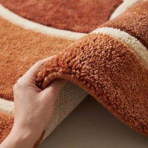 May include: Close-up of a plush, textured rug in shades of orange and cream. The rug features a geometric design with a soft, looped pile. The rug is being held up, revealing the underside.