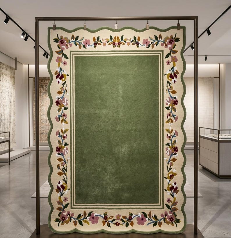 May include: A rectangular rug with a scalloped edge, featuring a solid olive green centre framed by a cream-coloured border. The border is decorated with a continuous floral vine pattern in shades of pink, yellow, burgundy, and green leaves.