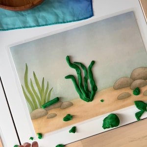 Puede incluir: Una escena de juego infantil con un fondo submarino impreso, algas marinas y rocas de plastilina verde. Piezas de plastilina verde dispersas realzan la escena. Un cuenco de madera y tela azul en el fondo sugieren una actividad creativa.