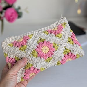 May include: A cream-colored crochet clutch with a floral pattern. The flowers are pink with yellow centers and green leaves. The clutch has a zipper closure and is held in a hand.
