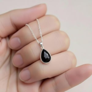 May include: A silver necklace with a black, teardrop-shaped gemstone pendant. The pendant is set in a silver bezel and suspended from a fine silver chain. The necklace is shown against a plain background.