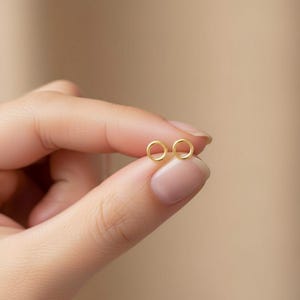 May include: A pair of small, gold-colored circle stud earrings. The earrings are held between fingers against a neutral background. The earrings are simple and minimalist in design, with a polished finish.
