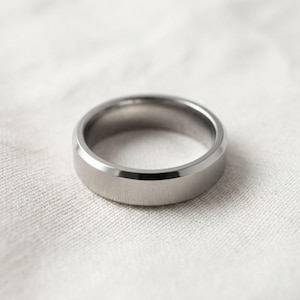 May include: A silver-colored ring with a flat top and beveled edges. The ring is made of a shiny metal and sits on a white, textured surface. The ring is a simple, classic design.