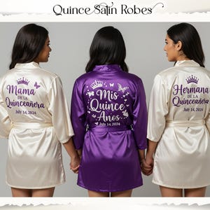 Peut inclure: Trois peignoirs en satin sont présentés, deux ivoire et un violet. Les peignoirs ivoire portent les inscriptions "Mama de la Quinceañera" et "Hermana de la Quinceañera" au dos, avec la date "July 14, 2024". Le peignoir violet indique "Mis Quince Años" avec la même date.