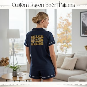 May include: Navy blue rayon short pyjama set with white trim. The back of the top features the text "Dreaming of Zero Deductions" in gold lettering. The pyjama set includes a collared, short-sleeved shirt and matching shorts.