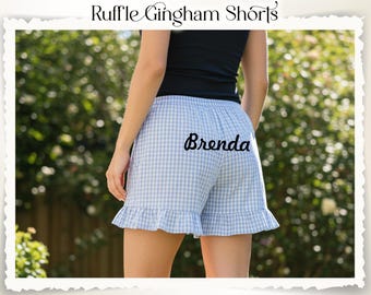 Puede incluir: Pantalones cortos de cuadros vichy azul claro y blanco con dobladillo con volantes. Los pantalones cortos tienen el nombre "Brenda" impreso en la parte posterior. La imagen también incluye el texto "Ruffle Gingham Shorts" en la parte superior.
