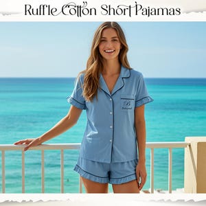 May include: Light blue short pajama set featuring a button-down shirt with a collar, short sleeves with ruffle trim, and matching shorts with ruffle trim. The shirt includes a chest pocket with the embroidered letter B and the text Bridesmaid.