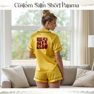 May include: Yellow satin short pyjama set with black trim. The pyjama top has short sleeves and the text "WAKE UP & HIT ZERO" in red. The set includes a button-down shirt and matching shorts.