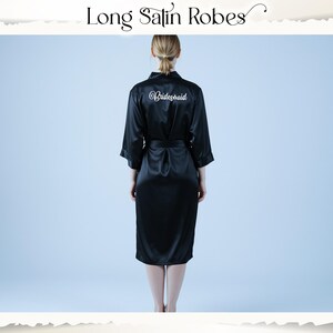 May include: A black satin robe with a matching belt. The word "Bridesmaid" is written in white script on the back. The robe is knee-length and has three-quarter sleeves. The text "Long Satin Robes" is at the top of the image.