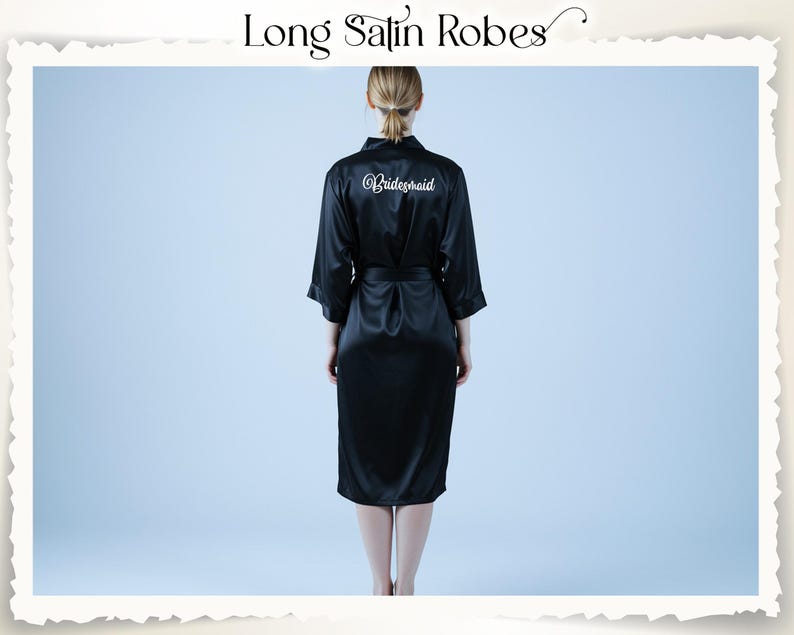 May include: A black satin robe with a matching belt. The word "Bridesmaid" is written in white script on the back. The robe is knee-length and has three-quarter sleeves. The text "Long Satin Robes" is at the top of the image.