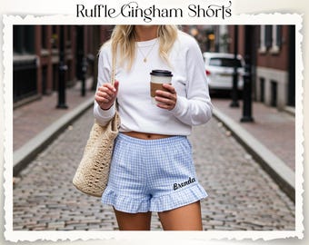 Puede incluir: Pantalones cortos de cuadros vichy azules y blancos con dobladillo con volantes. Los pantalones cortos tienen el nombre "Brenda" bordado en la parte delantera. La imagen también incluye el texto "Ruffle Gingham Shorts".