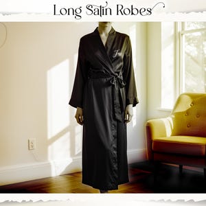 May include: A long black satin robe with a tie waist and long sleeves. The robe has the word "Jess" embroidered on the chest. The image also includes the text "Long Satin Robes". A yellow armchair is in the background.