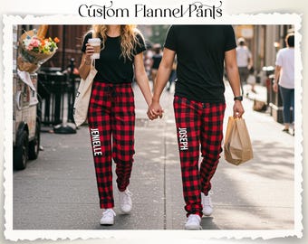 May include: Custom flannel pants in a red and black plaid pattern. The pants have the name "JENIELLE" printed vertically on one leg and "JOSEPH" on the other. The image shows a couple wearing the pants while walking down a city street.