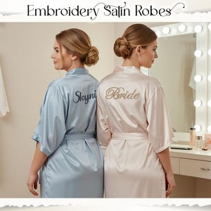 May include: Two women wearing satin robes. One robe is light blue with "Skyni" embroidered on the back, and the other is a pale pink with "Bride" embroidered on the back. The robes have a matching belt. The image is framed with a decorative border.