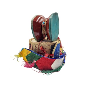 Puede incluir: Un tambor damaru tibetano tradicional con un acabado rojo y turquesa, apoyado sobre una cesta tejida. Una faja de tela colorida con borlas en rojo, amarillo, verde y azul está drapeada debajo del tambor.
