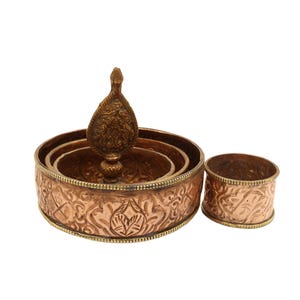 Op de afbeelding: Een decoratieve set van koperkleurig metaal met een sierlijk ontwerp. De set bevat een gelaagde kom met een decoratieve bekroning en een kleiner ringvormig stuk. De stukken hebben een gedetailleerd, reliëfpatroon.