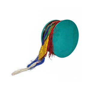 Puede incluir: Un tambor de mano turquesa con serpentinas de tela coloridas. El tambor tiene forma circular con una superficie verde azulado y está adornado con borlas rojas, amarillas, azules y blancas. El tambor está sobre un fondo blanco.