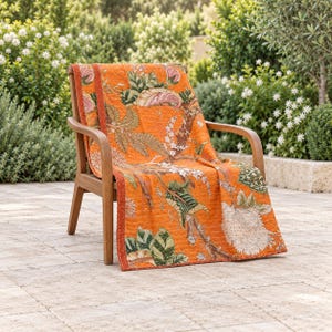 May include: A wooden armchair draped with an orange quilted throw blanket featuring a botanical print of large green leaves, brown branches, and white floral clusters. The blanket has visible stitching throughout and a solid orange border.