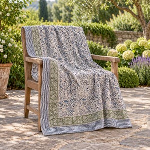 May include: A quilted throw blanket draped over a wooden chair, featuring a dense floral pattern in shades of blue and cream, bordered by a sage green floral band and a thin navy and white striped edge, creating a layered textile appearance.