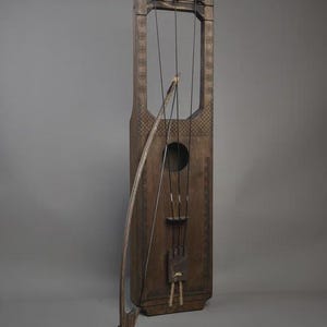 May include: A tall, dark brown wooden bowed string instrument with a rectangular body, a circular sound hole, and three vertical strings. The instrument features intricate geometric carvings along its edges and includes a matching wooden bow.