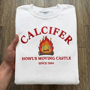 May include: White crew neck sweatshirt featuring red embroidered text "CALCIFER" and a cartoon flame character. Below the character, the text reads "HOWL'S MOVING CASTLE SINCE 2004".