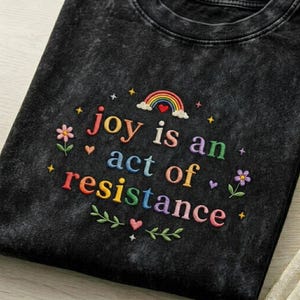 Può includere: T-shirt nero con testo ricamato "joy is an act of resistance" in lettere colorate. Il design include un arcobaleno, fiori, cuori e stelle. La maglietta ha uno scollo tondo.