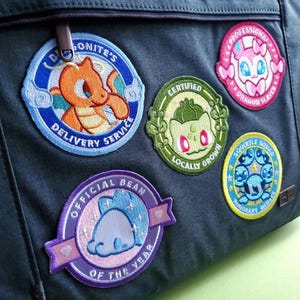Op de afbeelding: Een verzameling geborduurde patches met stripfiguren en tekst. De patches bevatten een Charmander met "Delivery Service", een Bulbasaur met "Locally Grown" en een Sylveon met "Dragon Slayer". Andere patches zijn "Official Bean of the Year" en "Squirtle Squad".