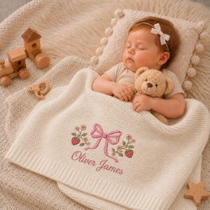 May include: A sleeping baby lies under a cream-coloured, embroidered blanket with a pink bow and floral design. The blanket is personalised with the name "Oliver James". A teddy bear is held by the baby. Wooden toys are nearby.
