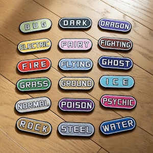 Puede incluir: Un conjunto de parches bordados con varias etiquetas de texto, incluyendo "BUG", "DARK" y "DRAGON". Cada parche tiene un fondo de color y un borde contrastante. Los parches están dispuestos sobre una superficie de madera.