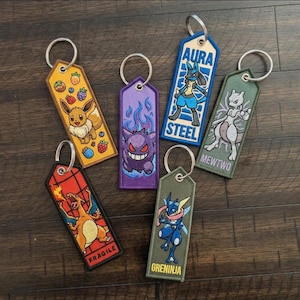 May include: Embroidered keychains featuring various Pokémon characters. The keychains have a rectangular shape with a metal ring. Designs include Eevee, Gengar, Lucario, Mewtwo, Charizard, and Greninja. Each keychain has a different color scheme and character.