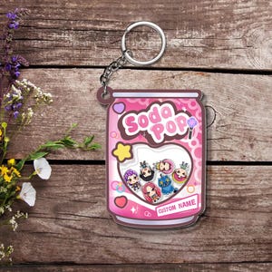 May include: A pink soda pop can-shaped keychain with a silver keyring. The keychain features the words "Soda Pop" in white, a heart-shaped window with cartoon characters, and the words "Custom Name".