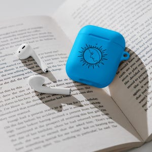 May include: A bright blue AirPods case with a sun and moon design, next to white wireless earbuds. The case is made of a smooth material and is open. The earbuds are resting on an open book.