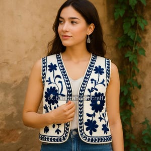 May include: A cream-coloured sleeveless vest featuring dark blue floral embroidery and trim. The vest is worn over a white top and blue jeans. The text "Chandrahandicraft" is visible on the vest.