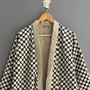 May include: A long-sleeved robe featuring a black fabric base with a repeating pattern of white circles. The garment includes a solid beige collar and front trim, two large front pockets, and a matching tie belt. The text Chandrahandicraft is visible.