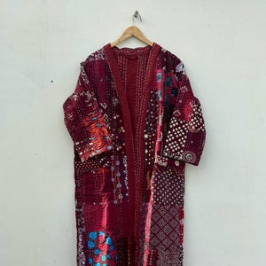 May include: A long, burgundy patchwork robe with a quilted design. The robe features various fabric patterns and colors, including blue, white, and orange. It is hanging on a wooden hanger against a plain white wall.