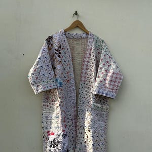 May include: A long, open-front robe with a patchwork design. The robe features a variety of floral and geometric patterns in shades of white, red, blue, and green. It has long sleeves and two front pockets, and is hanging on a wooden hanger.